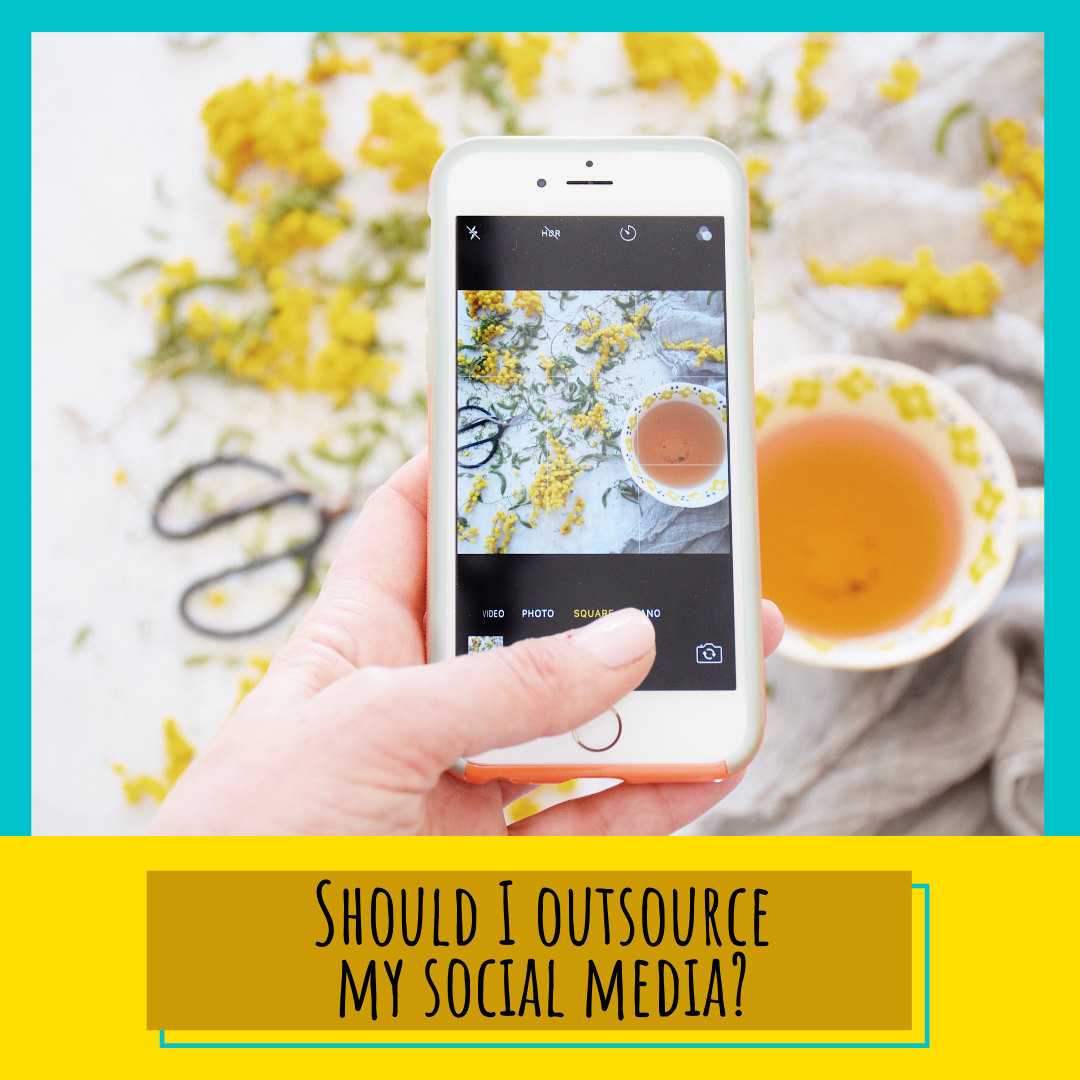 Hand holding a phone taking a picture of yellow flowers with text, "Should I outsource my social media?"