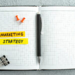 An open notebook, two thumb tacks, a pen and a clip are in the photo. Words written in the notebook are "marketing strategy".