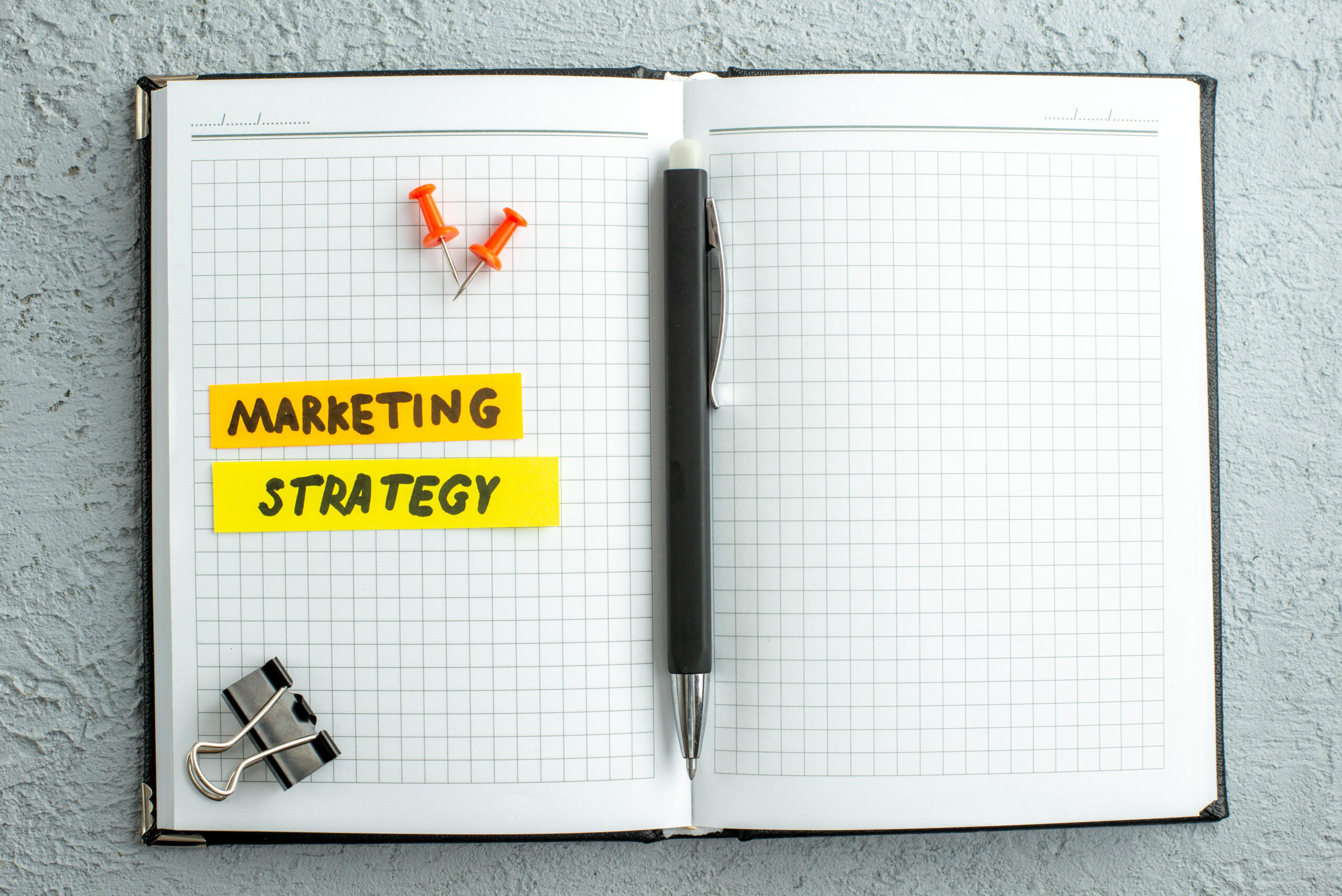 An open notebook, two thumb tacks, a pen and a clip are in the photo. Words written in the notebook are "marketing strategy".